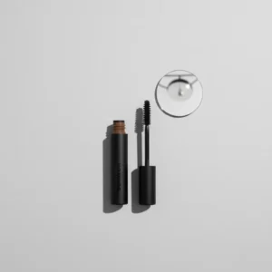 A minimalist flat lay composition on a light grey background, featuring an open tube of brown eyebrow mascara, its wand separated, and a small mirror. The style is clean, modern, and graphic, with sharp focus and even lighting