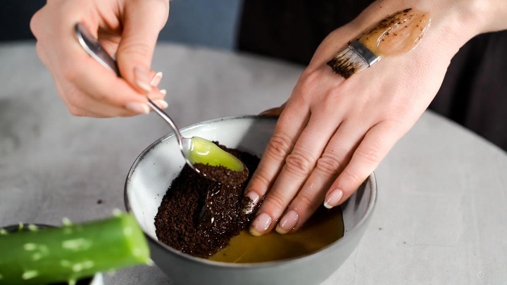 Close-up of a person's hands with clean, neutral manicured nails scooping and applying a creamy white aloe vera gel eye mask mixture from a gray ceramic bowl onto the back of one hand, demonstrating texture in a skincare swatch shot on a neutral background.